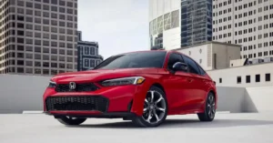 A red 2026 Honda Civic Si hatchback is parked on a rooftop with a cityscape in the background. The car is angled towards the viewer, with its front left side most prominent