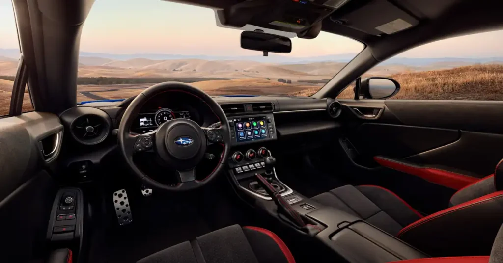 A wide-angle shot from inside a Subaru BRZ car shows the interior and the landscape outside. The car's interior is black with red accents on the seats and door panels. The steering wheel is black with red stitching and the Subaru logo in the center.