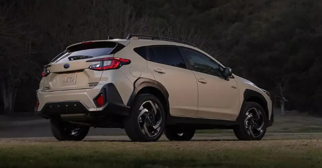 A tan 2026 Subaru Crosstrek Wilderness is parked on a grassy area with a dark, wooded background. The SUV is viewed from the rear right side, with its taillights and rear bumper clearly visible. The car has black trim around the wheel wells and a roof rack. The wheels are black with silver accents.