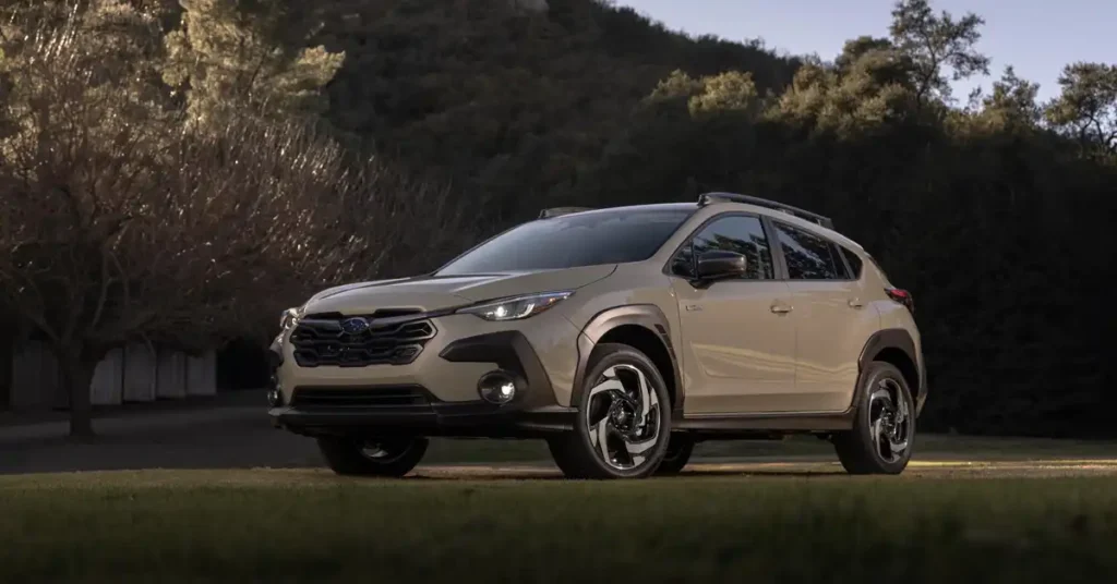 A tan 2026 Subaru Crosstrek is parked on a grassy area with a dark forest in the background. The car is facing left, with its front end slightly angled towards the viewer. The car has a black grille with the Subaru logo in the center, and sleek headlights.