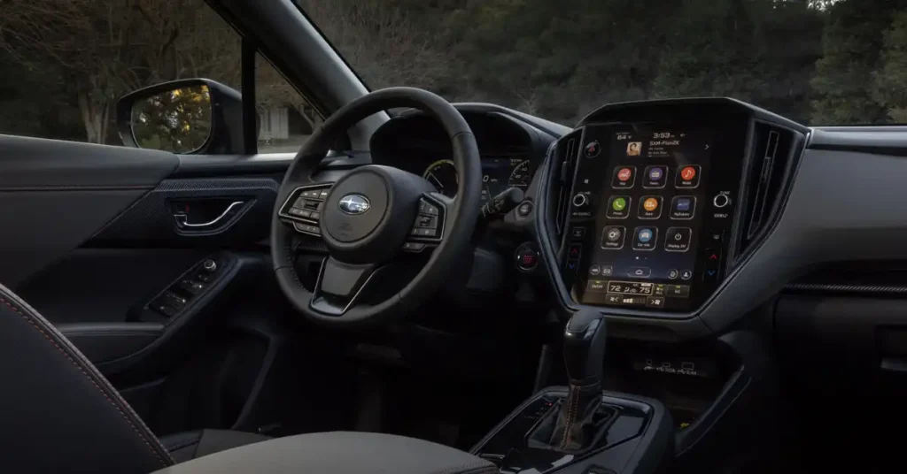 The interior of a2026 Subaru Crosstrekis shown from the perspective of the passenger seat. The image is dominated by dark tones, with black leather and dark gray accents. In the foreground, the edge of a dark gray seat with red stitching is visible. To the left, the car door panel features black leather, a textured carbon fiber-like insert around the door handle, and a row of black buttons for window controls.