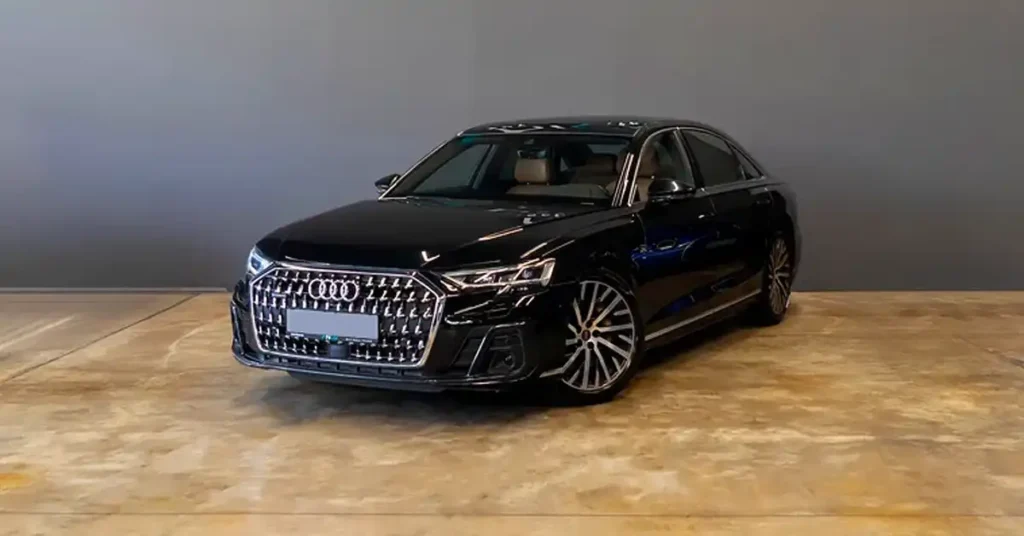 A black Audi A8 sedan is parked on a concrete floor in front of a gray wall. The car is facing the viewer at a slight angle, with its front grille and headlights prominently displayed. The grille is large and chrome-plated, with a distinctive honeycomb pattern.