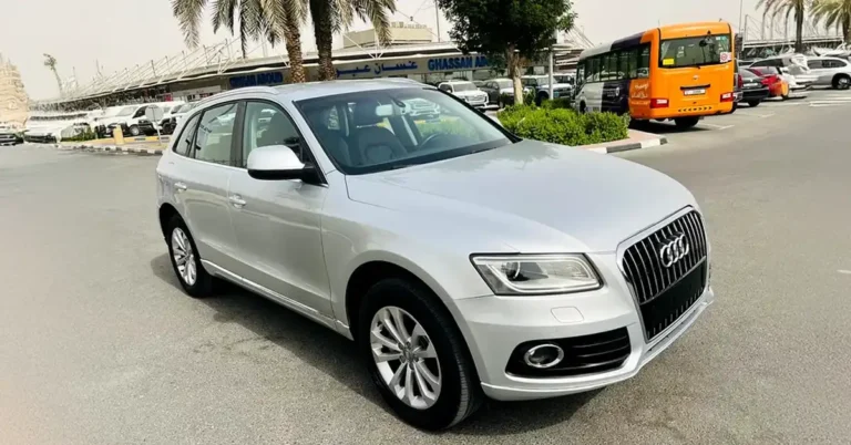 A silver Audi Q5 SUV is parked in an outdoor lot. The car is facing the viewer at a slight angle, with its front left side most prominent. The vehicle has a sleek, modern design with chrome accents on the grille and fog lights. The tires are black with silver alloy wheels.