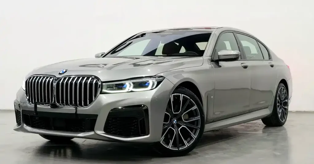 A silver BMW 7 Series sedan is shown from a low-angle, front-three-quarter view. The car is parked on a polished concrete floor in a studio with a white background. The vehicle has a prominent chrome grille with vertical slats and a BMW logo at the top.
