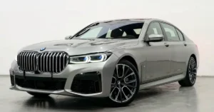 A silver BMW 7 Series sedan is shown from a low-angle, front-three-quarter view. The car is parked on a polished concrete floor in a studio with a white background. The vehicle has a prominent chrome grille with vertical slats and a BMW logo at the top.