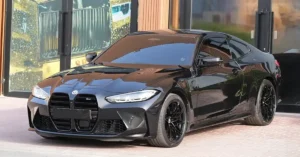 A black BMW M4 coupe is parked in front of a building with large glass windows and wooden paneling. The car has a sleek, sporty design with sharp lines and a low profile. Its black paint is highly reflective, mirroring the surroundings.