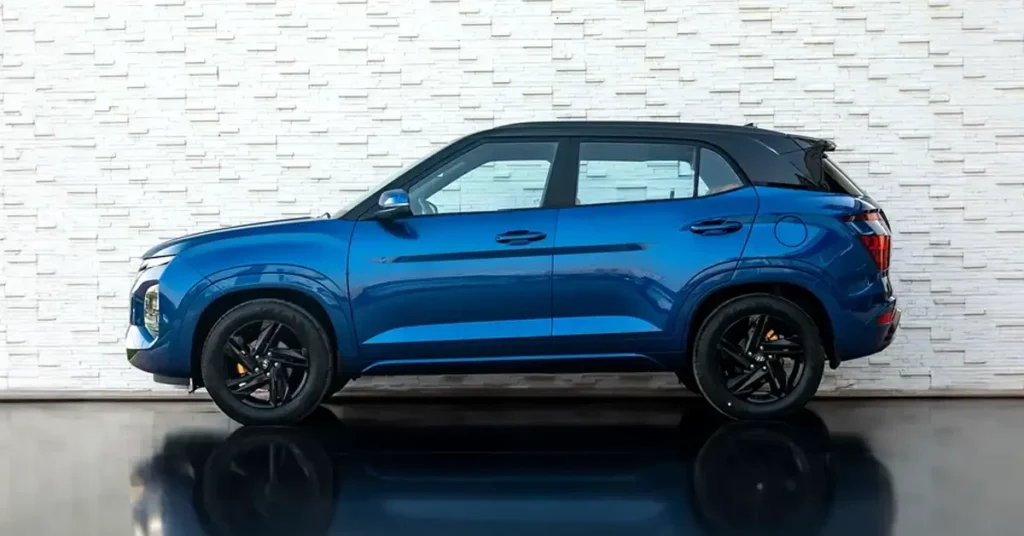 A side view of a blue Hyundai Creta SUV with a black roof and black wheels, parked in front of a white brick wall. The car is facing left, and its reflection is visible on the shiny floor.