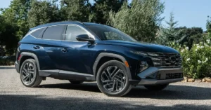 A side view of a dark blue 2024 Hyundai Tucson SUV, parked on a gravel road, with trees and bushes in the background. The car has a modern design with sharp lines and a distinctive front grille.