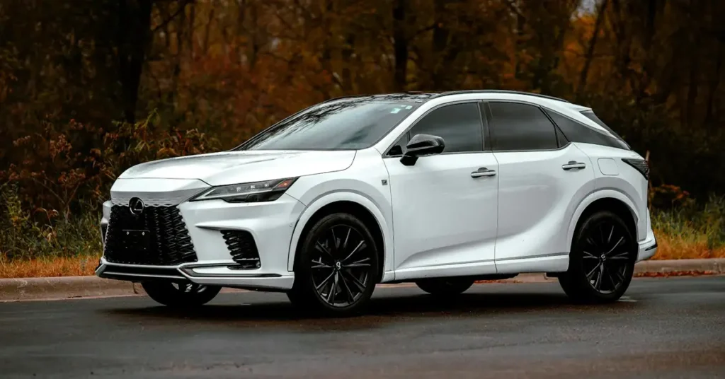 A white Lexus RX 500h F Sport Performance SUV is parked on a wet asphalt road, with a backdrop of trees showing fall colors. The car features a black roof, black rims, and tinted windows. The lighting is soft, highlighting the car's sleek design and the contrast between its white body and black accents.