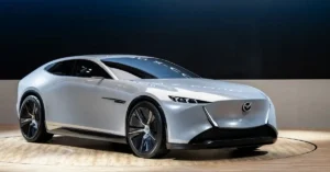 A Mazda Vision X Coupe, a sleek, futuristic Mazda coupe, is displayed on a rotating wooden platform. The car is a pearlescent white with a low profile and sharp, angular lines. Its front features slim, horizontal LED headlights and a prominent Mazda logo.