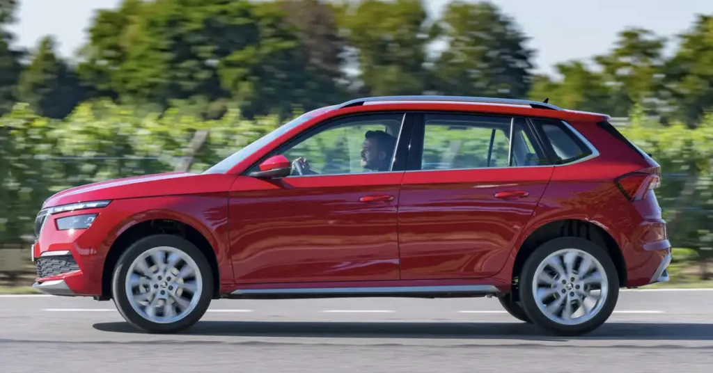 A side profile of a red Skoda Kamiq SUV is captured in motion on a paved road, with a blurred background of green trees and a bright sky. The car's metallic red paint gleams under the daylight, reflecting the surrounding environment.