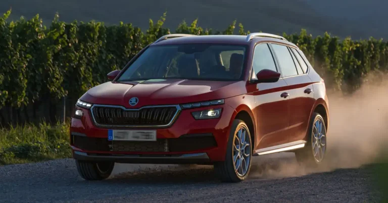 A red Skoda Kamiq SUV is driving on a dirt road, kicking up dust. The car is in motion, with its wheels turned slightly to the left. The sun is setting, casting a warm glow on the scene. The background is a vineyard with lush green vines.
