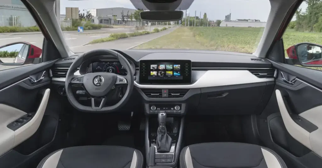 A wide-angle shot captures the interior of a Skoda Kamiq SUV, looking forward through the windshield. The dashboard is dominated by a large, central touchscreen displaying a grid of nine colorful icons. To the left, a digital instrument cluster shows a speedometer with a needle pointing to zero and other vehicle information.