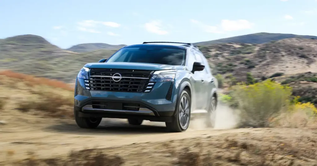 A teal Nissan Pathfinder SUV is driving on a dirt road in a dry, hilly landscape. The SUV is angled towards the viewer, with its front right wheel kicking up a cloud of dust. The vehicle has a black grille with a chrome Nissan logo, sleek headlights, and a roof rack. The background features rolling hills covered in dry brush and scattered green bushes, with a clear blue sky above.