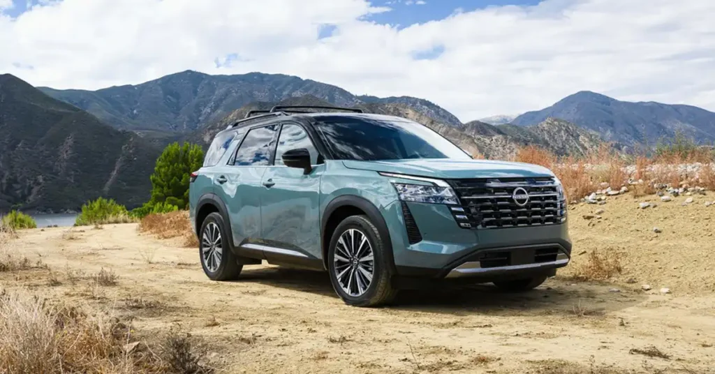 A teal 2026 Nissan Pathfinder SUV is parked on a dirt road with a mountainous landscape in the background. The SUV is facing the camera at a slight angle, with its front left side most prominent. It has a black roof rack and black trim around the wheel wells and windows.