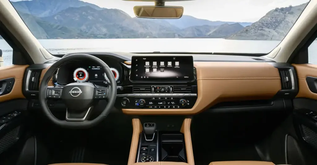 A wide-angle, eye-level shot shows the interior of a modern Nissan Pathfinder SUV, with a scenic view of a lake and mountains visible through the windshield. The dashboard is a blend of black and tan leather, with prominent stitching on the tan sections.