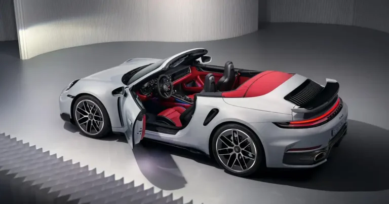 A silver 2026 Porsche 911 Turbo S convertible is parked in a studio with a light gray, ribbed wall in the background. The car's doors are open, revealing a luxurious red leather interior with black accents. The convertible top is down, exposing the black seats and the red interior lining.