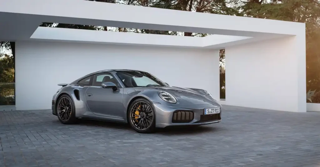 A silver Porsche 911 Turbo S is parked on a grey paved driveway in front of a modern white building with an overhang. The car is viewed from the front-left quarter, with its headlights on. The building has a minimalist design with clean lines and a large opening in the overhang, revealing trees and foliage in the background.