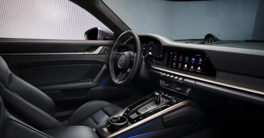 A digital rendering of the interior of a Porsche 911 Turbo S, is presented from the perspective of the passenger seat. The interior is predominantly black, with accents of brushed metal and carbon fiber. The seats are upholstered in black leather with visible stitching.