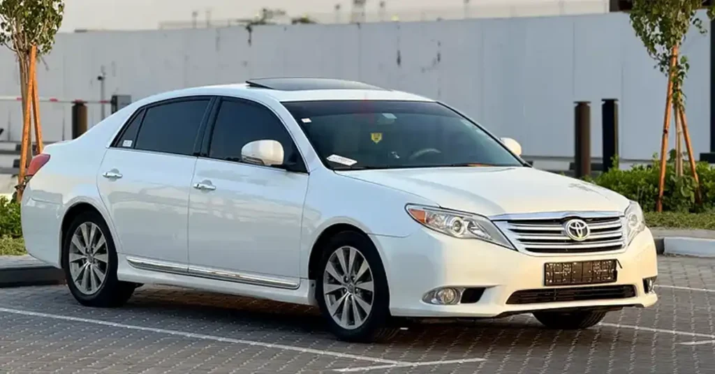 A black Toyota Avalon sedan is parked on a brick-patterned surface. The car has tinted windows and a sunroof. The interior appears to be red leather. The headlights are on, illuminating the front of the car