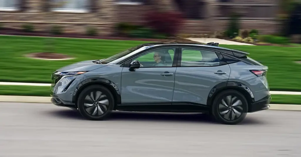 A side profile of a gray Nissan Ariya electric SUV is captured in motion, driving on a paved road. The car is sleek and modern, with a black roof and black wheel arches.