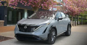 A light gray2025 Nissan Ariya Platinum+ e-4ORCE is parked on a paved surface in front of a modern building with large glass doors. The car is positioned at an angle, with its front facing the viewer. A tree with abundant pink blossoms is in the foreground, partially obscuring the top of the car and the building.