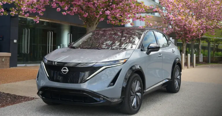 A light gray2025 Nissan Ariya Platinum+ e-4ORCE is parked on a paved surface in front of a modern building with large glass doors. The car is positioned at an angle, with its front facing the viewer. A tree with abundant pink blossoms is in the foreground, partially obscuring the top of the car and the building.