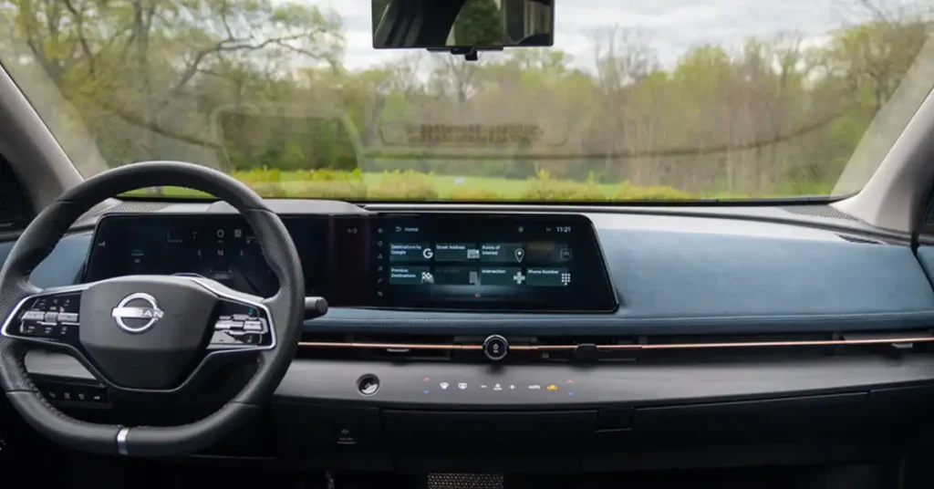 A wide shot captures the interior of a modern Nissan Ariya electric SUV, focusing on the dashboard and steering wheel. The steering wheel, on the left, is black with silver accents and features the Nissan logo prominently in the center. To the right of the steering wheel, a sleek, dark dashboard stretches across, housing two large, integrated digital screens. The left screen displays driving information