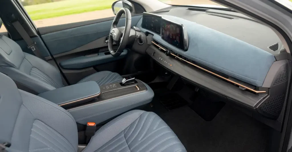 The image shows the interior of a 2025 Nissan Ariya Platinum+ e-4ORCE , focusing on the front passenger and driver's seats and dashboard. The seats are upholstered in a light blue, perforated leather with intricate stitching patterns. The center console features a sleek gear shifter and a padded armrest, also in the same blue leather with subtle metallic trim.