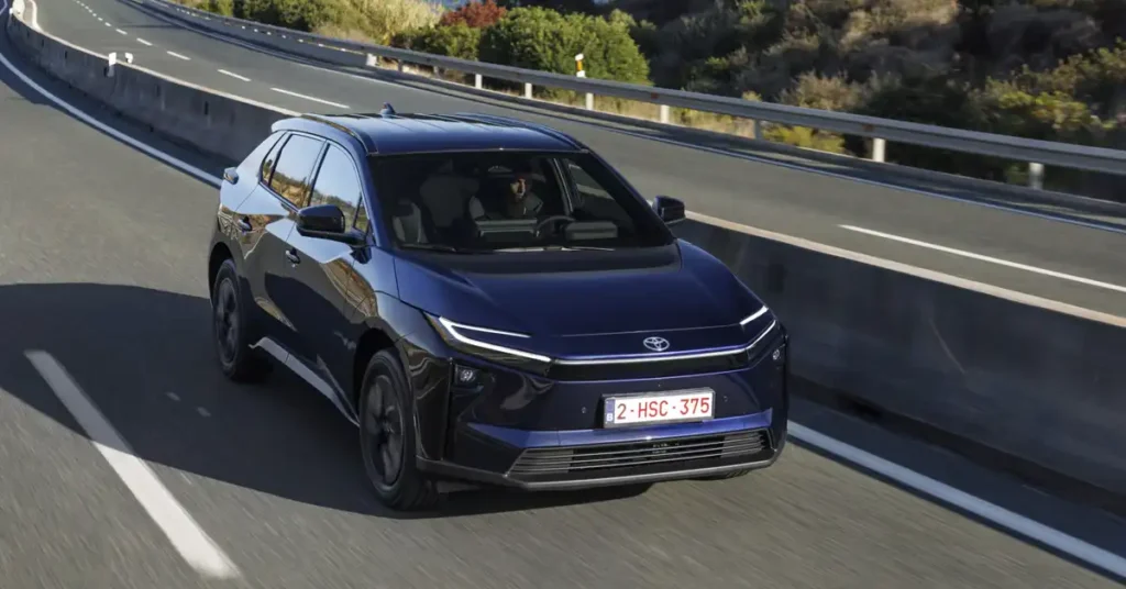 A dark blue 2025 Toyota bZ4X SUV is captured in a dynamic, low-angle shot as it drives on a highway. The car is positioned slightly to the left of the frame, with its front facing the viewer. The highway has multiple lanes marked with white lines, and a concrete barrier runs along the right side.