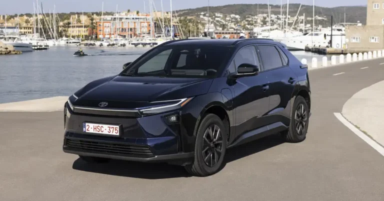 A dark blue 2025 Toyota bZ4X SUV is parked on a paved road next to a body of water. The SUV is in the foreground, facing slightly to the left. In the background, there is a harbor filled with sailboats and yachts, with buildings and hills visible beyond. The sky is clear and blue.