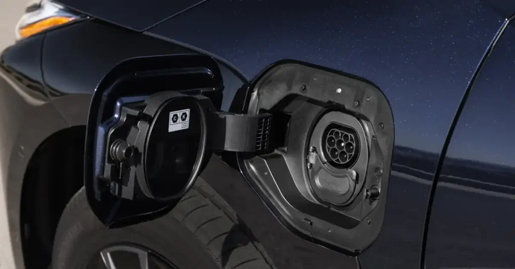 A close-up, eye-level shot shows the open charging port of a 2025 Toyota bZ4X SUV. The charging port cover is open, revealing the charging socket with its circular arrangement of pins.