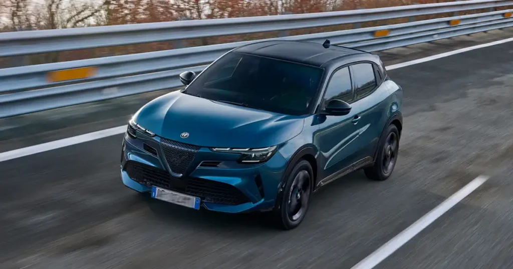 A dynamic, eye-level shot captures a sleek, teal-colored Alfa Romeo Junior Ibrida Q4 driving on a highway. The car, with its black roof and tinted windows, is positioned slightly to the left of the frame, its front end angled towards the viewer
