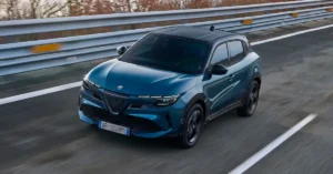 A dynamic, eye-level shot captures a sleek, teal-colored Alfa Romeo Junior Ibrida Q4 driving on a highway. The car, with its black roof and tinted windows, is positioned slightly to the left of the frame, its front end angled towards the viewer