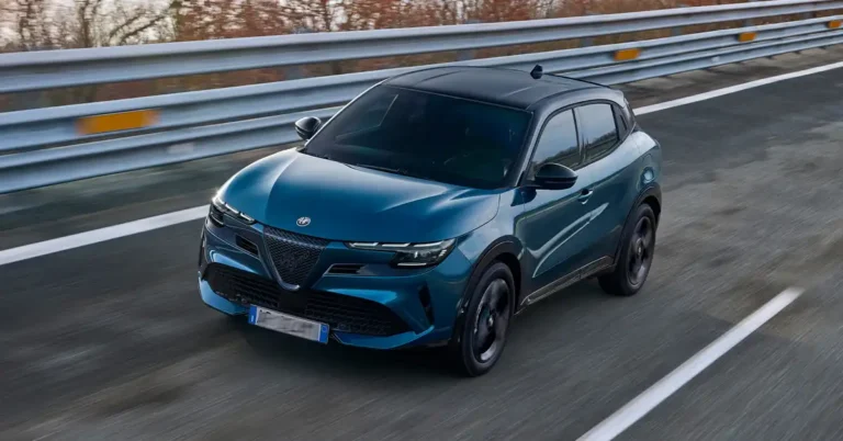 A dynamic, eye-level shot captures a sleek, teal-colored Alfa Romeo Junior Ibrida Q4 driving on a highway. The car, with its black roof and tinted windows, is positioned slightly to the left of the frame, its front end angled towards the viewer