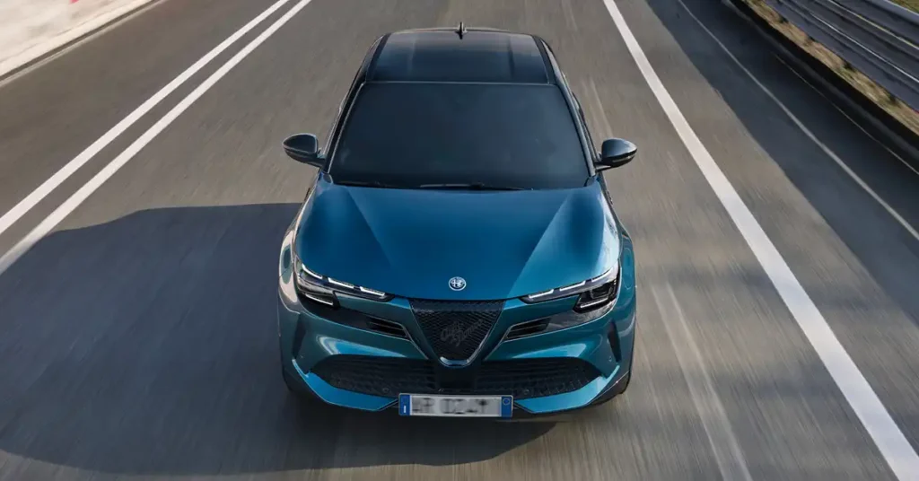 A high-angle, full shot shows a teal Alfa Romeo Junior Ibrida Q4 driving on a highway. The car is centered in the frame, with its front facing the viewer. The road has two white lines on either side of the car, indicating it is in its lane. The background is blurred, suggesting motion.