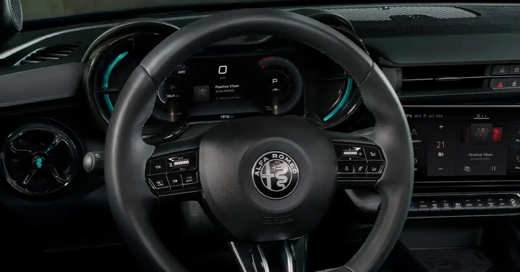 A close-up, eye-level shot of a Alfa Romeo Junior Ibrida Q4 interior focuses on the steering wheel and dashboard. The steering wheel, predominantly black leather, features the Alfa Romeo logo prominently in its center. Buttons for various functions are visible on the wheel's spokes.