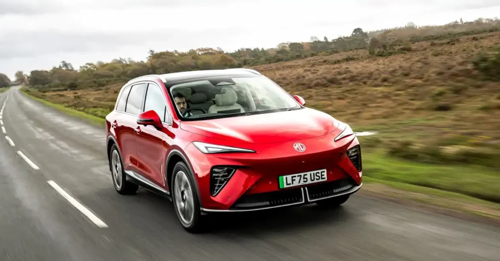 A dynamic, eye-level, medium shot captures a shiny redMG MGS6 EV SUV driving on a wet, asphalt road. The car is angled towards the viewer, with its front right wheel prominently displayed. A man with a beard is visible driving the car.