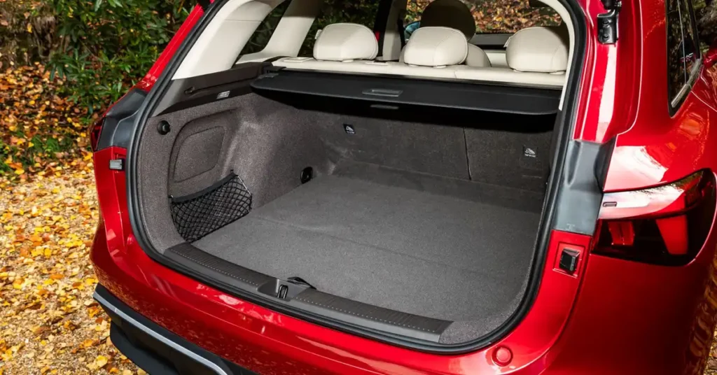 A close-up, eye-level shot shows the open trunk of a shiny MG MGS6 EV SUV. The trunk is empty and clean, with a gray carpeted floor and dark gray fabric lining the sides. A black cargo net is attached to the left side of the trunk.
