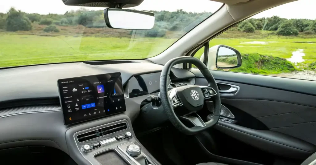 The image shows the interior of MG MGS6 EV SUV, focusing on the dashboard and steering wheel. A large touchscreen display is prominently featured on the left, showing various icons and information, including weather and navigation. To the right of the screen is a digital instrument cluster.