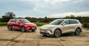 Two MG MGS6 EV SUVs, one red and one beige, are parked on a concrete surface with tire marks. The red SUV is on the left, slightly behind the beige SUV, which is on the right and closer to the viewer. Both vehicles are facing forward, with their front ends angled slightly towards the camera