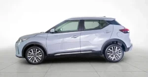 A light gray Nissan Kicks is shown in a studio setting against a white background. The car is viewed from the side, with its front on the left and rear on the right. The vehicle has black trim around the wheel wells and along the bottom of the doors.