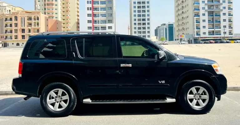 A black Nissan Armada SUV is parked on a paved surface in front of a row of modern buildings. The SUV is viewed from the side, with its front half cut off by the frame.