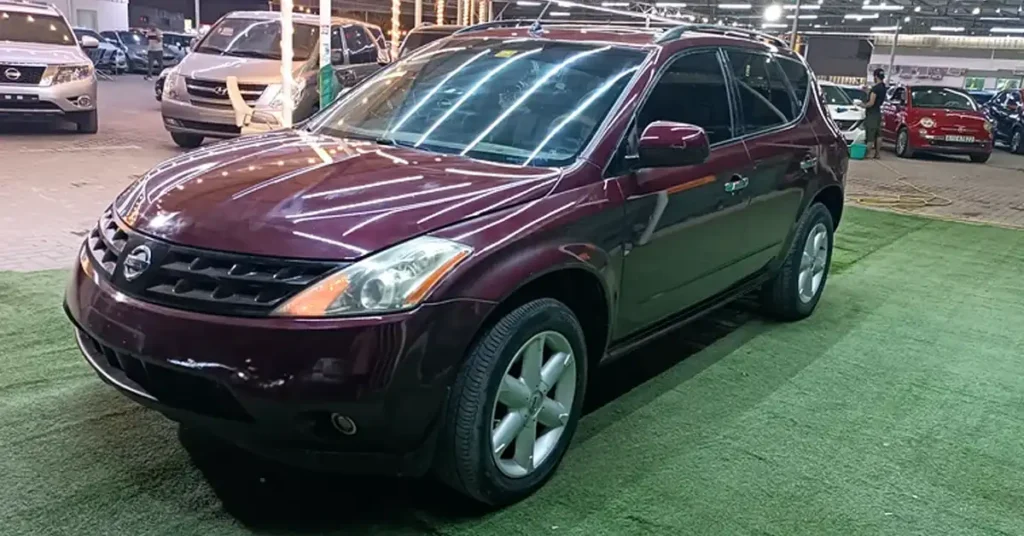 A maroon Nissan Murano SUV is parked on a green artificial turf in an indoor parking lot. The car is angled towards the viewer, with its front left side most prominent. The vehicle's headlights and grille are visible, as is its shiny paint reflecting the overhead fluorescent lights.