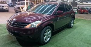 A maroon Nissan Murano SUV is parked on a green artificial turf in an indoor parking lot. The car is angled towards the viewer, with its front left side most prominent. The vehicle's headlights and grille are visible, as is its shiny paint reflecting the overhead fluorescent lights.