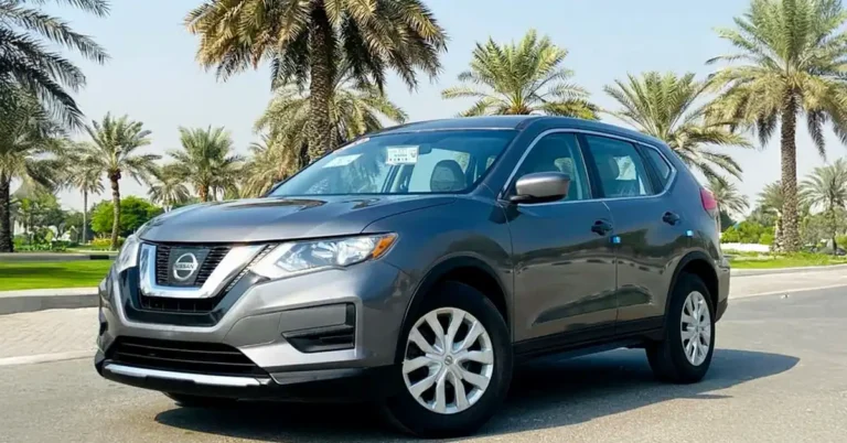 A dark gray Nissan Rogue SUV is parked on the side of a road, with its front end angled towards the viewer. The car is in focus, with the background blurred. Behind the car, several palm trees with lush green fronds are visible against a clear blue sky.