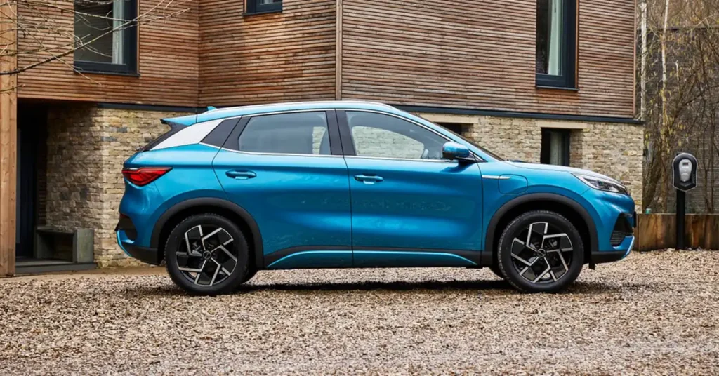 A blue electric BYD ATTO 3 SUV is parked on a gravel driveway in front of a modern house with wooden and stone accents. The car is shown in profile, facing right, with its doors closed.