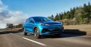 A blue 2025 BYD ATTO 3 electric SUV is driving on a paved road. The car is in the foreground, and the background consists of trees and a blue sky with some clouds. The road has white lines on the side, and the car is casting a shadow on the road