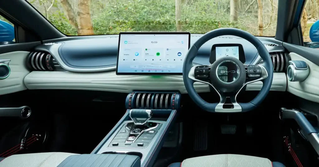 A wide shot shows the interior of a BYD ATTO 3 , focusing on the dashboard and steering wheel. The dashboard is a light cream color with blue accents and features a large, central touchscreen displaying a clean interface with various app icons.