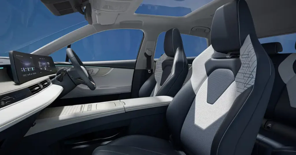 A wide-angle shot of the interior of a 2025 Chery OMODA E5 Interior Experience and Digital Comfortmodern car, looking forward from the passenger side. The focus is on the front seats, which are upholstered in a two-tone design of dark gray and light gray with a geometric pattern on the backrests.